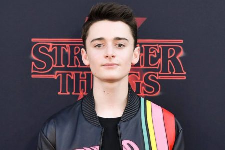 Foto do ator Noah Schnapp. Ele é branco, cabelo preto com topete e usa um casaco preto com listras coloridas - Metrópoles