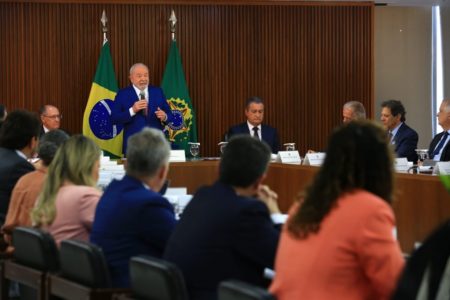 O presidente Lula faz primeira reunião ministerial no Palácio do Planalto e pede boa relação com o Congresso Nacional. Na imagem ele está de pé, na ponta de uma mesa, falando em microfone com bandeiras do Brasil atrás e ao lado de Alckmin - Metrópoles