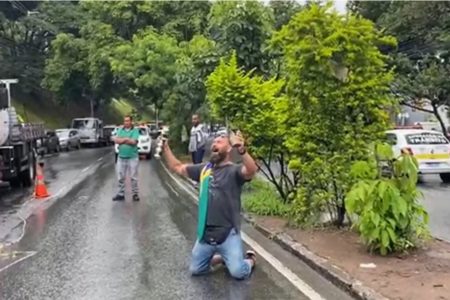 Bolsonarista chora pelo fim de acampamento na frente do QG do Exército em Belo Horizente no meio da pista. Ele ergue as mãos ao céu com bandeira do Brasil no ombro - Metrópoles