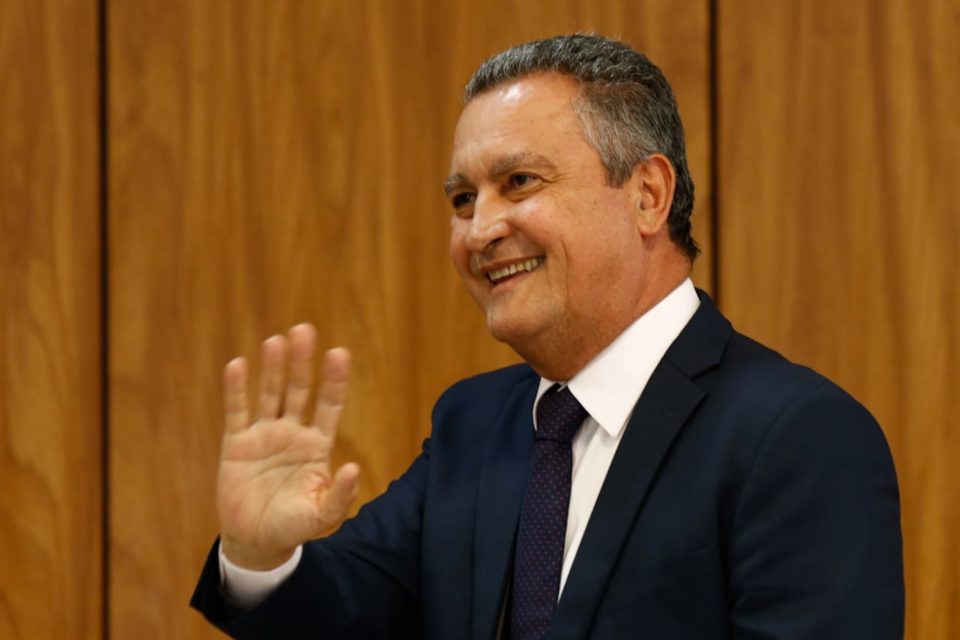 Ministro da Casa Civil, Rui Costa, concede entrevista coletiva sobre primeira reunião ministerial do governo Lula. Ele acena para a plateia e sorri, sentado diante de mesa - Metrópoles