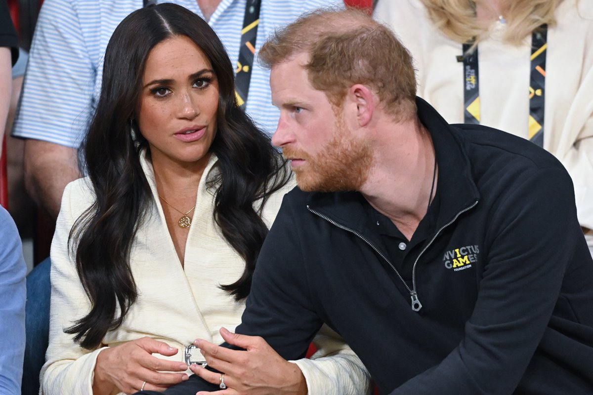 A atriz norte-americana Meghan Markle ao lado do seu marido, o príncipe Harry da família real britânica, em uma arquibancada durante um evento esportivo. Ela, uma mulher negra com cabelo liso longo, usa um casaco de manga longa branco. Ele, branco com cabelo curto ruivo, usa um casaco preto com um zíper na gola. - Metrópoles