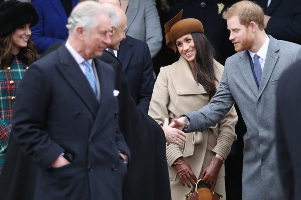 O princípe Harry, um homem branco e jovem, de cabelo liso ruivo, ao lado da esposa, a atriz Meghan Markle. Ela é uma mulher jovem negra, de cabelo castanho longo. Eles cumprimentam pessoas que passam pela rua. Na frente do casal está o rei Charles, um homem velho e branco, de cabelos curtos brancos. - Metrópoles