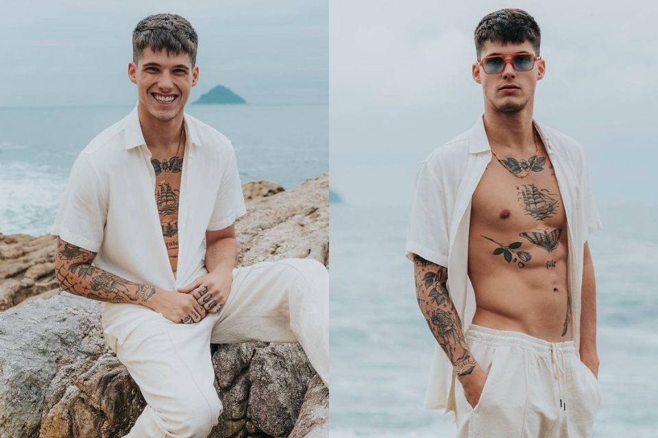 Gabriel, participante da Casa de Vidro do BBB23, em fotos posadas na praia. Ele sorri usando uma camisa branca aberta e calça, exibindo suas tatuagens ao longo do torso e braço - Metrópoles