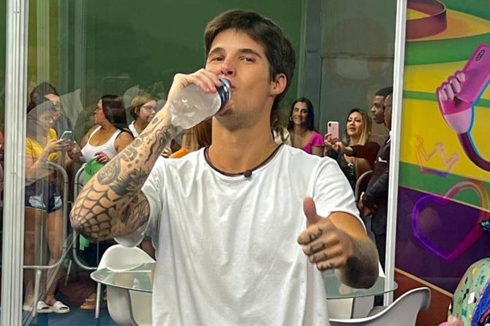 Gabriel, participante da Casa de Vidro do BBB23. Ele aparece dentro do confinamento em shopping, com público filmando atrás, e dá sinal de joinha enquanto bebe água - Metrópoles