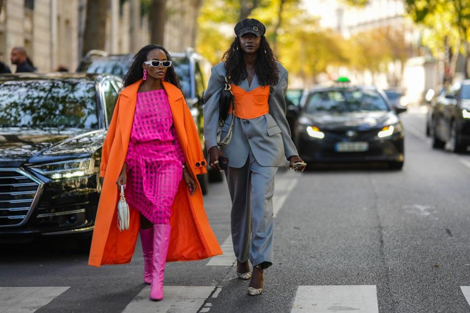 Street style: em faixa de pedestre, amigas caminham lado a lado. Uma delas usa look rosa com sobretudo laranja. A outra veste conjunto cinza de alfaiataria com sobreposição de corset laranja - Metrópoles