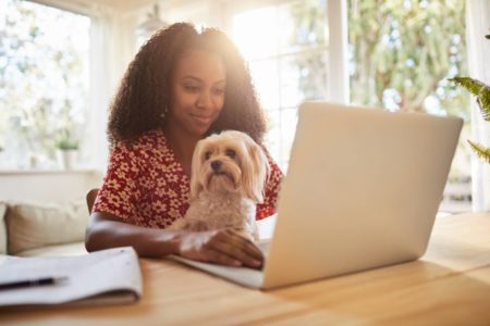 Moça sentada na frente de um notebook enquanto segura um cachorro muito fofinho no colo