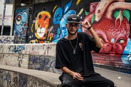 Foto colorida do rapper Canaan. Ele usa boné preto, camisa preta e calça preta. Está sentado em uma calçada - Metrópoles