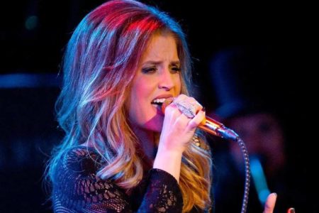 Lisa Marie Presley está utilizando anéis em sua mão direita, segura um microne e canta. Ela é branca e tem os cabelos claros - Metrópoles