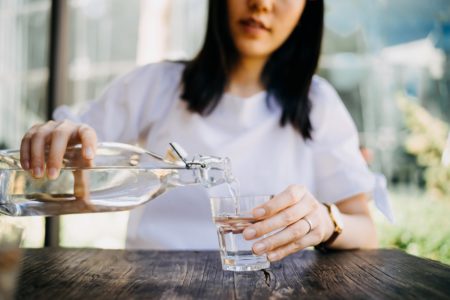 Mulher branca e jovem, de cabelos lisos pretos, enchendo um copo de vidro de água. Com a mão direita ela segura a garrafa. - Metrópoles