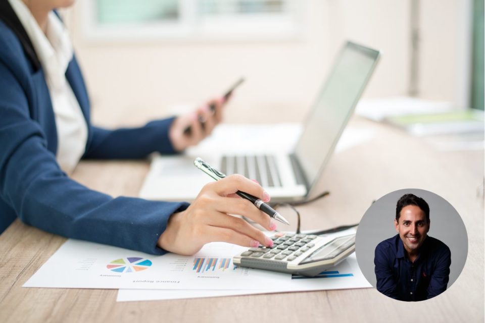 Uma pessoa branca com terno azul está com uma mão na calculadora e a outra no celular. A mesa ainda é composta por notebook e papéis. No canto direito tem um círculo com a foto do especialista em finanças Charlles Di