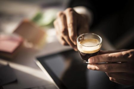 Homem negro segurando uma pequena xícara de café em cima de um tablet. o fundo, aparece uma mesa com papéis e caneta, mostrando que eles está em um escritório