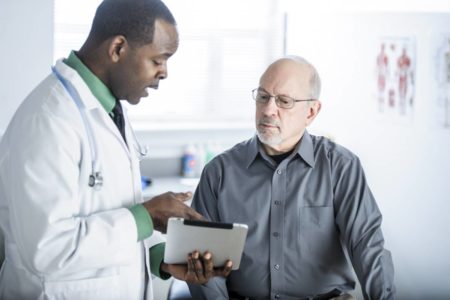 Médico segurando pranhcete de diagnóstico e explciando a situação para paciente idoso que usa óculos