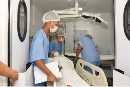 Imagem colorida de uma sala de cirurgia com pessoas vestindo roupas hospitalares azuis e mascara