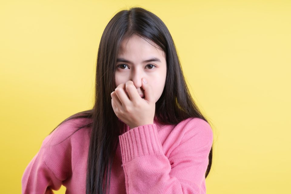 Uma menina branca, com cabelos pretos lisos está tapando o nariz como se algo tivesse com odor ruim. O fundo da imagem é amarelo e ela está com um casaco rosa