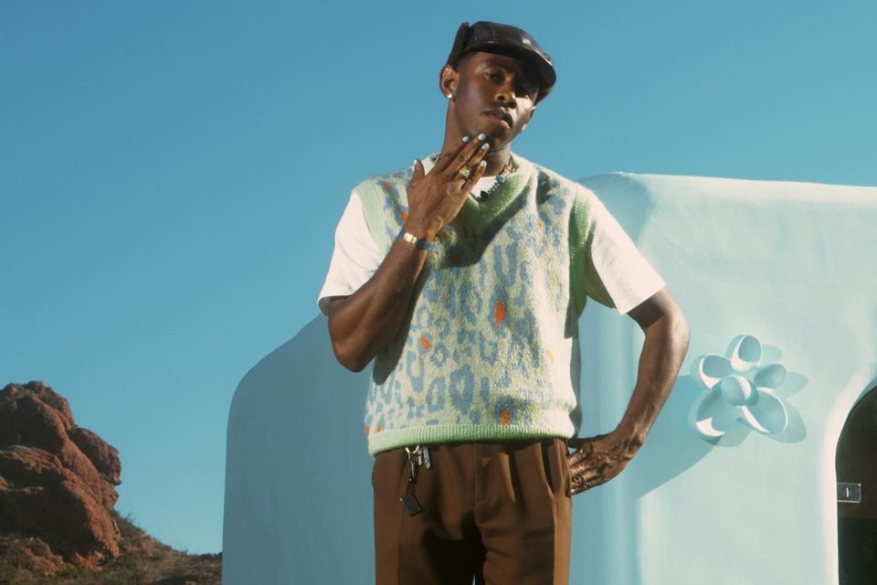 O rapper Tyler, The Creator, um homem negro e jovem, de cabelos raspados. Na imagem, ele usa uma camiseta branca, um colete verde colorido, uma calça marrom e uma boina cinza. - Metrópoles