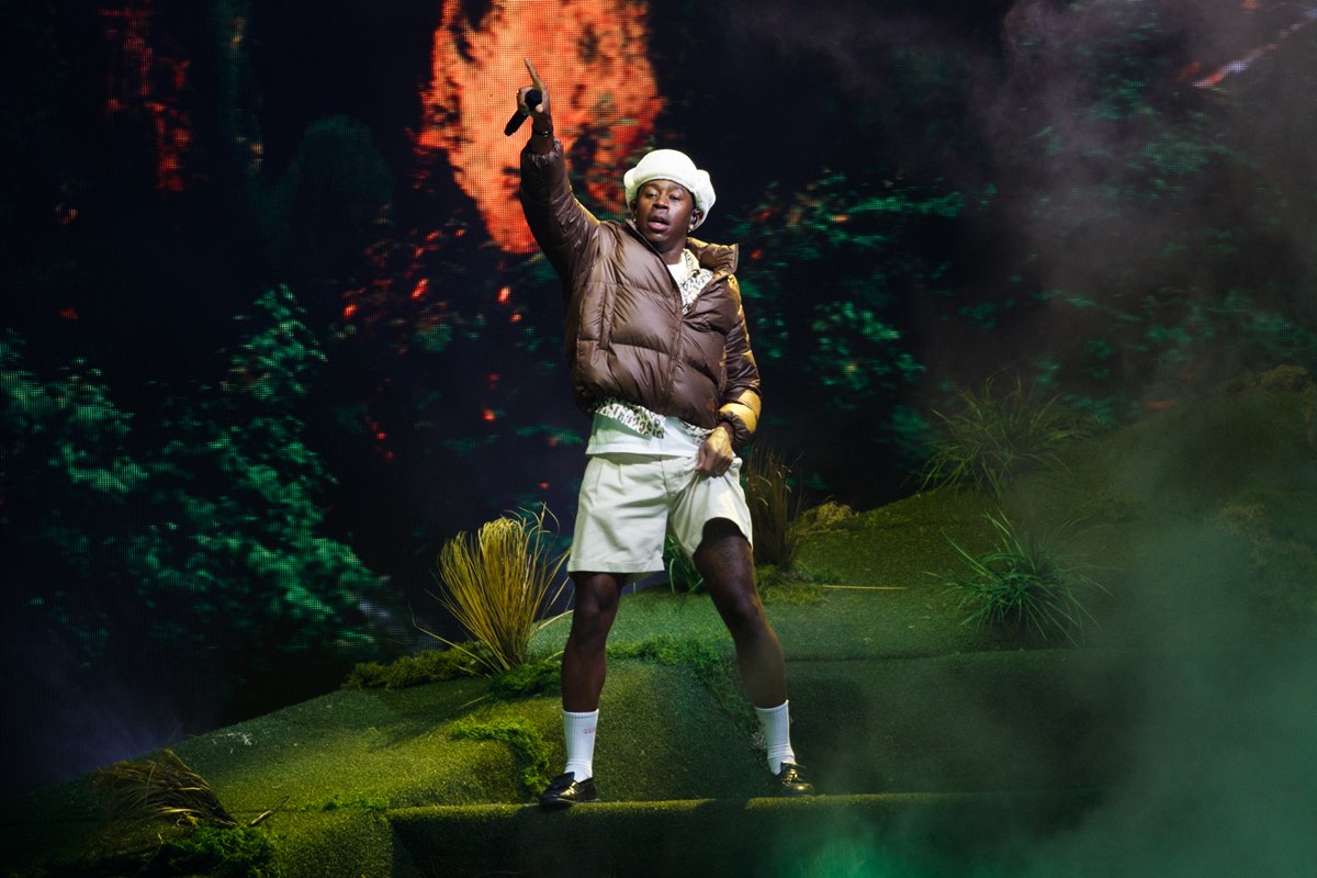 O rapper Tyler, The Creator, um homem negro e jovem, de cabelos raspados, cantandon o palco de um show. Na imagem, ele usa um conjunto de camiseta e bermuda, ambos em um tom de verde limão claro, um casaco marrom e um chapéu verde de peludo. - Metrópoles