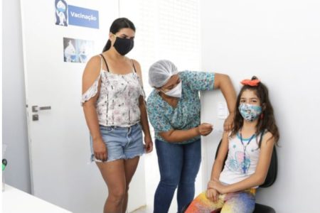 Imagem colorida de três pessoas: duas mulheres e uma criança. Uma das adultas está aplicando uma vacina no braço da criança