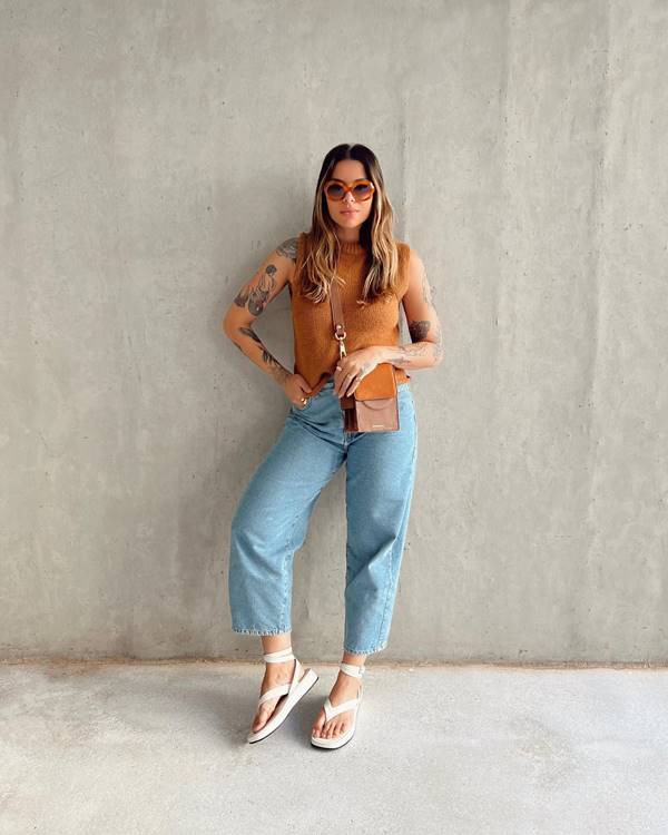 Mulher encostada em parede usa calça jeans com camiseta bege e óculos grande - Metrópoles