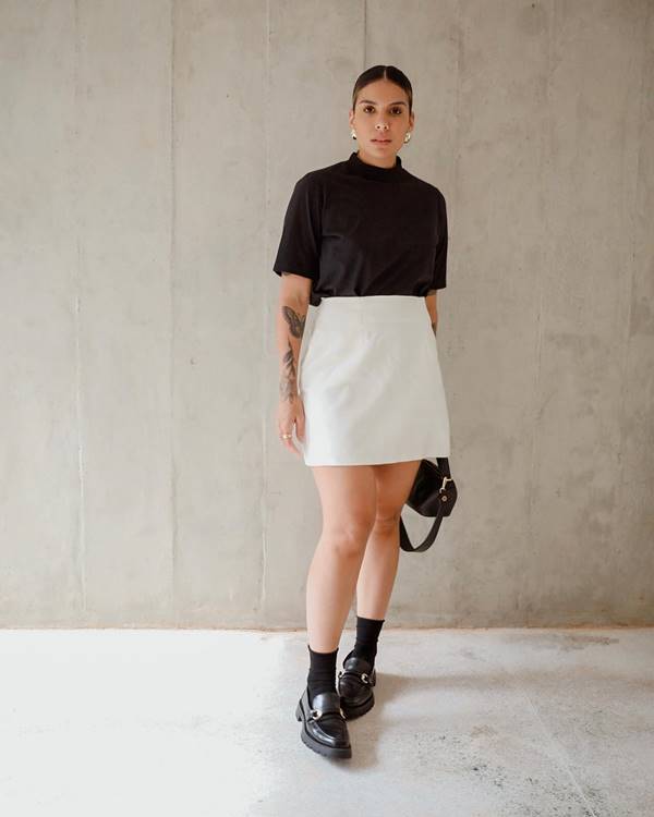 Mulher usa camiseta preta com modelagem gola, saia branca, meia preta e mocassim - Metrópoles