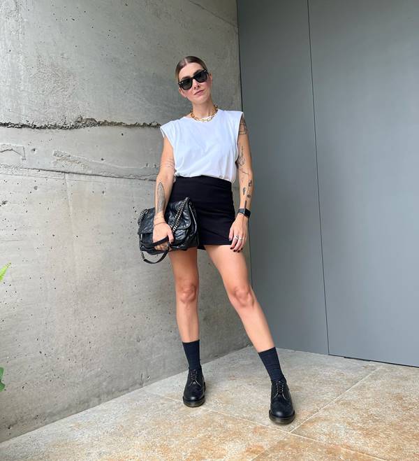 Mulher branca com cabelo preso usa camiseta branca, short preto, meia preta de cano alto e mocassim - Metrópoles