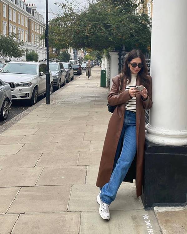 mulher branca encostada na parede usa calça jeans e sobretudo na cor marrom - Metrópoles 