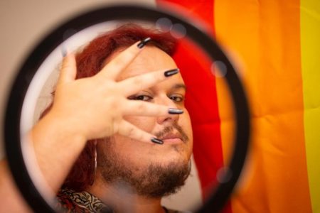 Pessoa com cabelo vermelho e barba mostrando a mão com unhas pretas em frente ao rosto. Atrás dele há uma bandeira com as cores vermelha e laranja