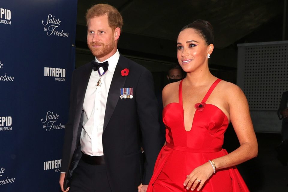 Na imagem, Harry e Meghan estão de mãos dadas em evento de gala. Ele está usando um terno preto e ela está com o cabelo amarrado em coque e usando um vestido todo vermelho - Metrópoles