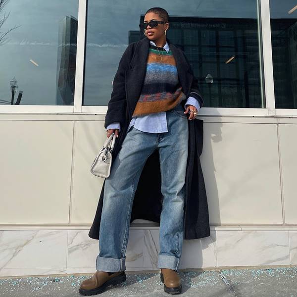 Influencer negra usa colete com listras em cores neutras, casaco preto sobretudo, calça jeans larga e bota - Metrópoles