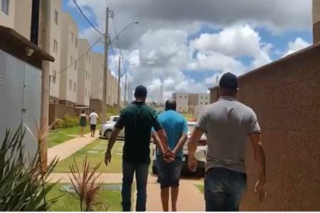 Imagem colorida de três homens. Um deles segura a mão do outro que está amarrada.