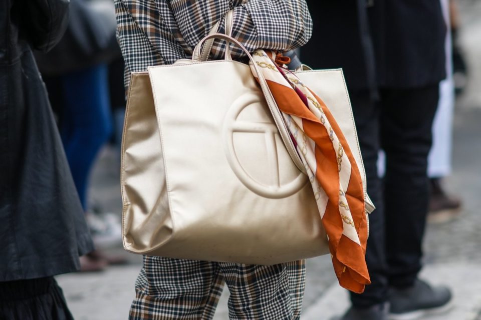 Bolsa quadrada e grande, de couro dourado, da marca Telfar. A pessoa que a segura amarrou um lenço marrom na alça da bolsa. - Metrópoles