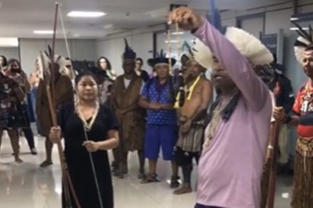 Indígenas Pataxó fazem ritual de "limpeza espiritual" no Ministério da Saúde. Na imagem, em primeiro plano, um líder segura colar e mulher ao lado segura arco e flecha enquanto outros membros da tribo, caracterizados, observam em corredor - Metrópoles