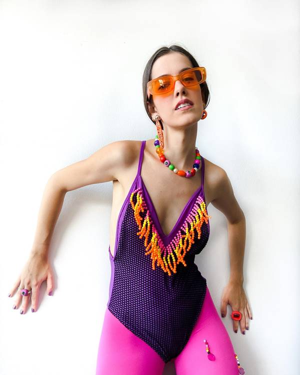 Modelo usa regata roxa, colar de miganças e cinto de miçangas. O visual dela é completado com um óculos de sol de lente laranja e luvas douradas - Metrópoles