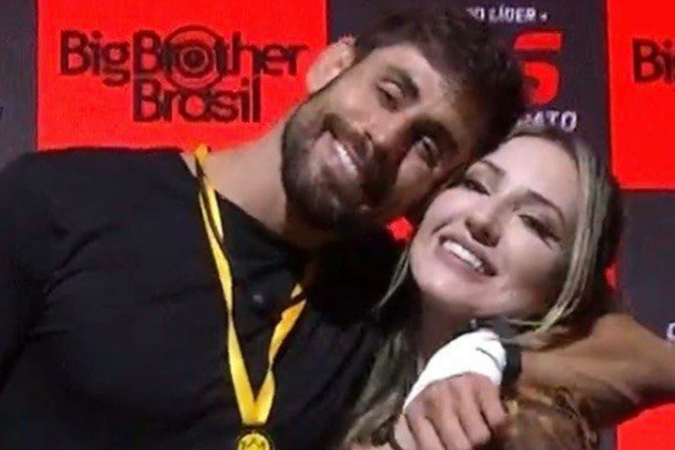 Cara de Sapato e Amanda. Ele está com um colar amarelo e roupa preta. Ela está de roupa vermelha - Metrópoles