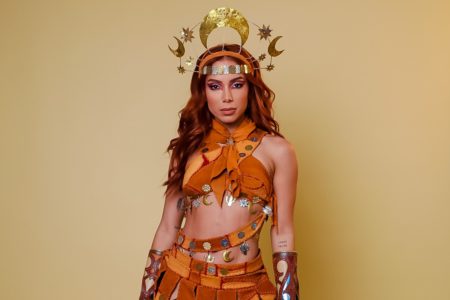 A cantora Anitta, uma mulher parda e jovem, de cabelo ondulado longo e acobreado, posa para foto em um fundo bege liso. Ela usa um top e uma saia de couro, além de uma tiara tourada. A roupa faz uma referência ao uniforme do cangaço usado por Maria Bonita. - Metrópoles