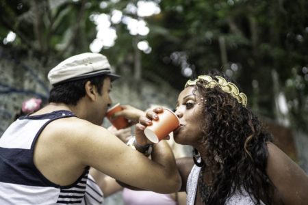 Imagem de um homem e uma mulher fantasiados para o Carnaval, cruzando os braços e bebendo de um copo colorido - Metrópoles