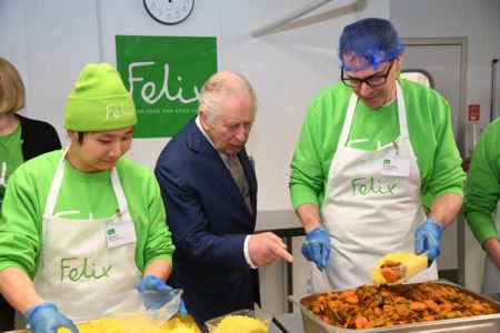 Foto colorida de homem idoso, branco e vestido com terno apontando para comida. Ele está entre dois homens vestidos com roupas verdes e um avental bege - Metrópoles