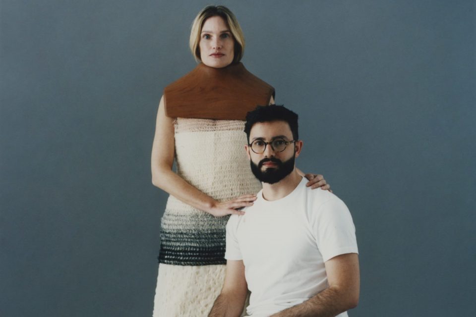 O estilista João Maraschin, ao lado de uma modelo, posa para foto em um fundo liso cinza. Ele é um homem branco e jovem, de cabelo e barba preta, e usa uma camisa branca. A modelo é uma mulher branca de meia idade, cabelo liso loiro, e usa um top de crochê. - Metrópoles