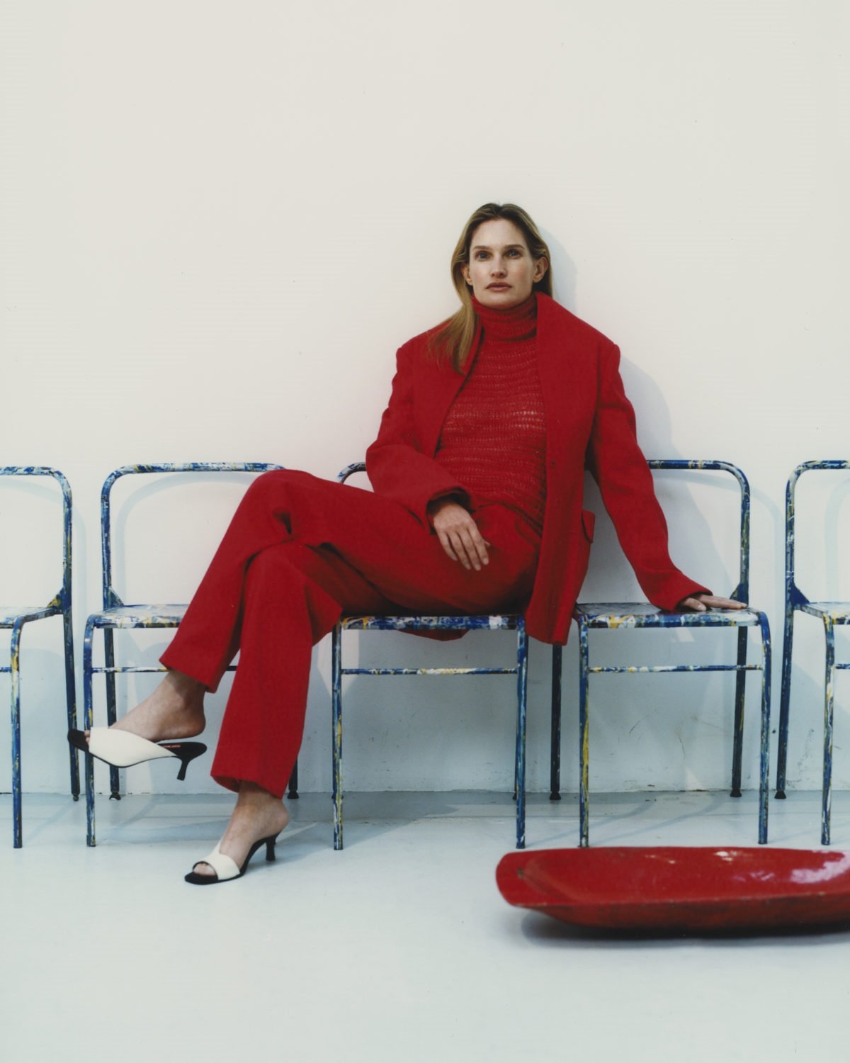 Fotos de campanha da marca do estilista João Maraschin. Na imagem, uma modelo branca e de meia idade, de cabelo liso loiro, está sentada em uma cadeira de metal em um fundo branco. Ela usa um conjunto de crochê vermelho composto por blusa, calça e casaco. - Metrópoles