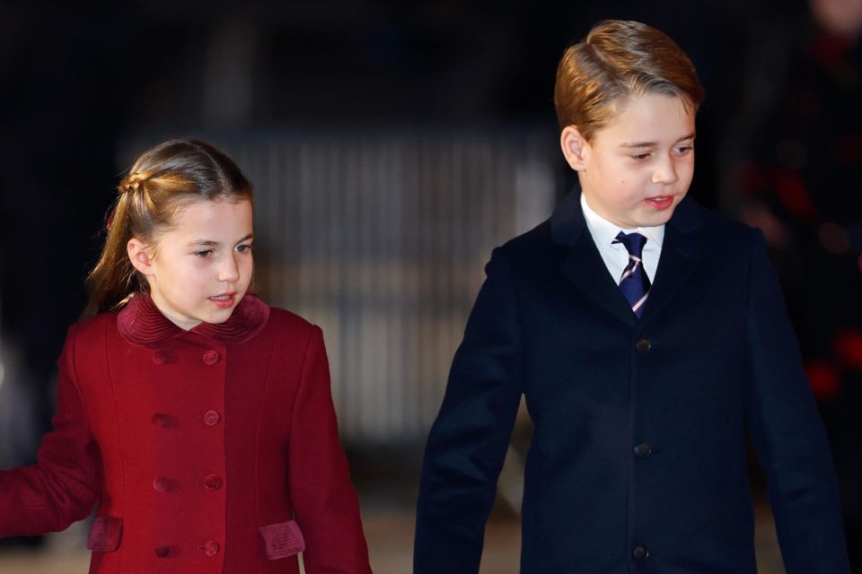 Foto colorida de menina branca, loira e com casaco vermelho ao lado de menino branco, loiro, vestido com um casaco preto. Ele usa gravata azul e blusa branca - Metrópoles