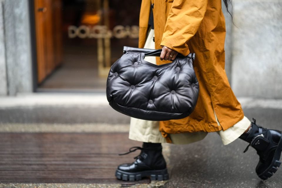 Mulher andando na rua segurando bolsa preta. Ela está usando casaco mostarda e sapatos pretos - Metrópoles