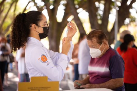 Mulher com jaleco segurando seringa com vacina e idoso ao fundo em ambiente com árvores