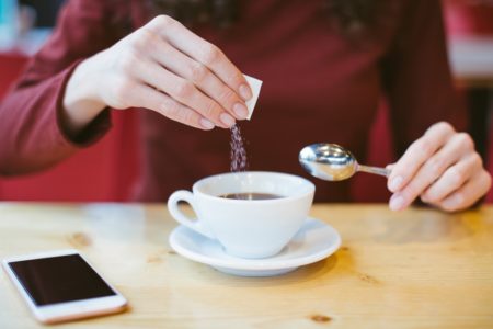 Foto colorida de mão branca colocando sachê de adoçante em xícara de café. Ao lado, há um celular e a outra mão segura uma colher pequena - Metrópoles