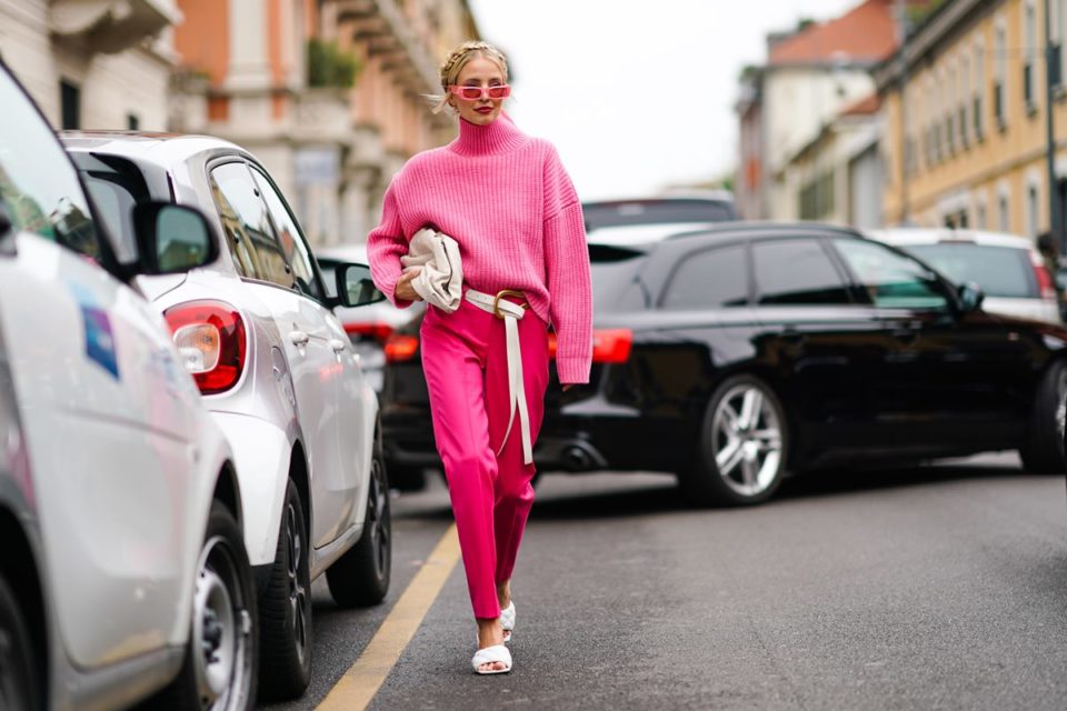 Na rua, mulher entre carros usa look rosa com calça de alfaiataria e suéter com gola alta rosa, além de sandália branca. Ela segura uma bolsa branca - Metrópoles