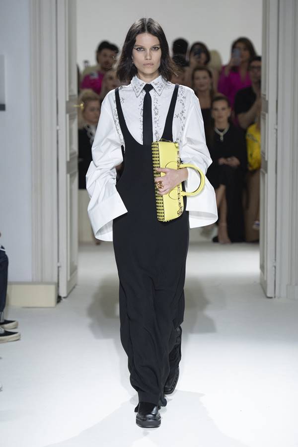 Modelo branca usa macacão preto , camisa branca, gravata preta e segura bolsa amarela no braço - Metrópoles