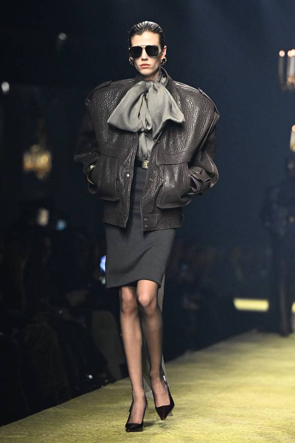 Modelo usa saia lápis preta, casaco de couro preto e blusa de gola com laço grande - Metrópoles