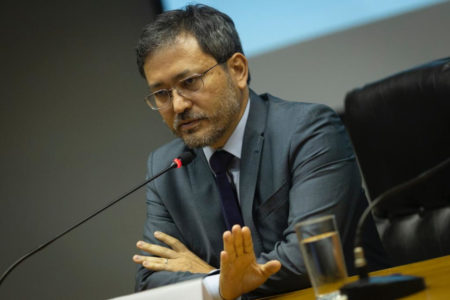 homem de terno sentado a mesa com microfone a frente gesticula