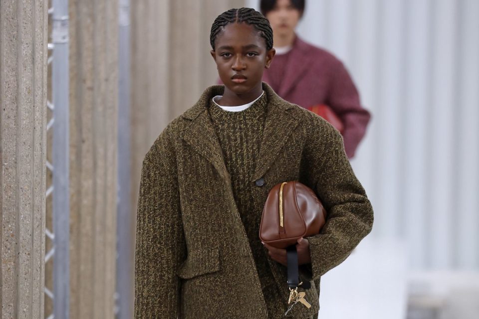 adolescente Zaya Wade, uma mulher trans e negra, de cabelos trançados pretos, no desfile da Miu Miu, na Semana de Moda de Paris. Ela usa um vestido e um casaco, ambos na cor verde, e segura uma bolsa de couro marrom. - Metrópoles