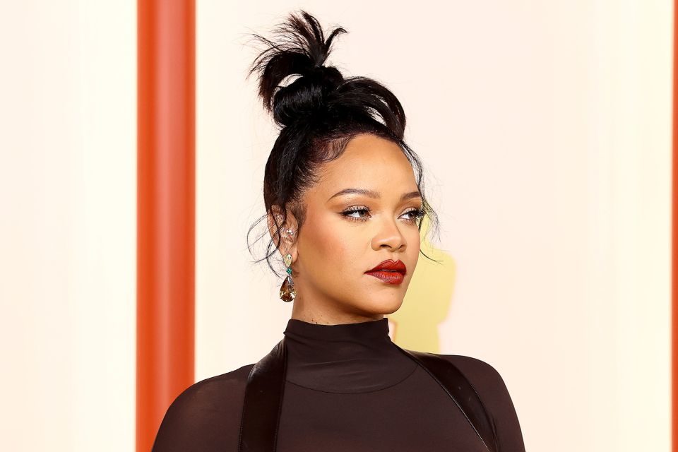 Foto da cantora Rihanna. Ela está de coque, batom vermelho e look preto - Metrópoles