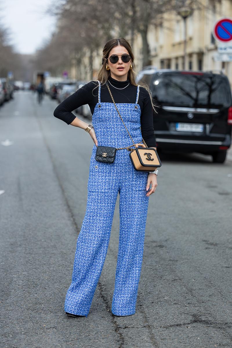 Mulher branca e jovem, de cabelo liso castanho, posa para foto na rua durante a Semana de Moda de Paris. Ela usa um top preto de manga longa e gola alta, um macacão de tweed azul, uma blusa marrom de palha e uma pochete de couro, tudo da marca Chanel. - Metrópoles