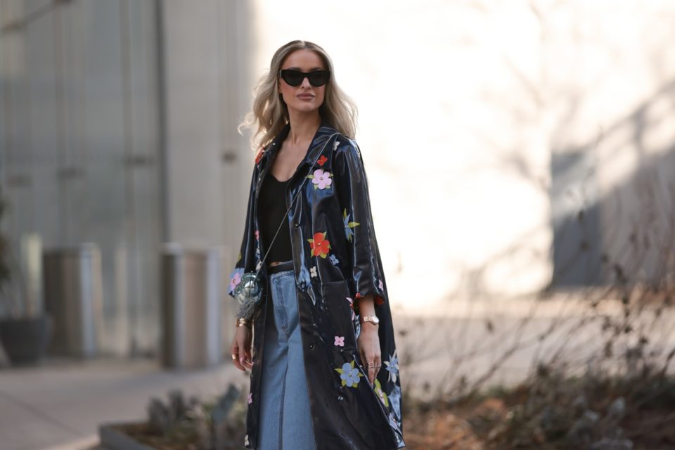 Mulher branca e jovem, de cabelo loiro e onduldo, posa para foto andando nas ruas de Nova York. Ela usa um top preto, calça jeans, óculos escuros e uma jaqueta de chuva preta, cintilante, com flores bordadas. - Metrópoles